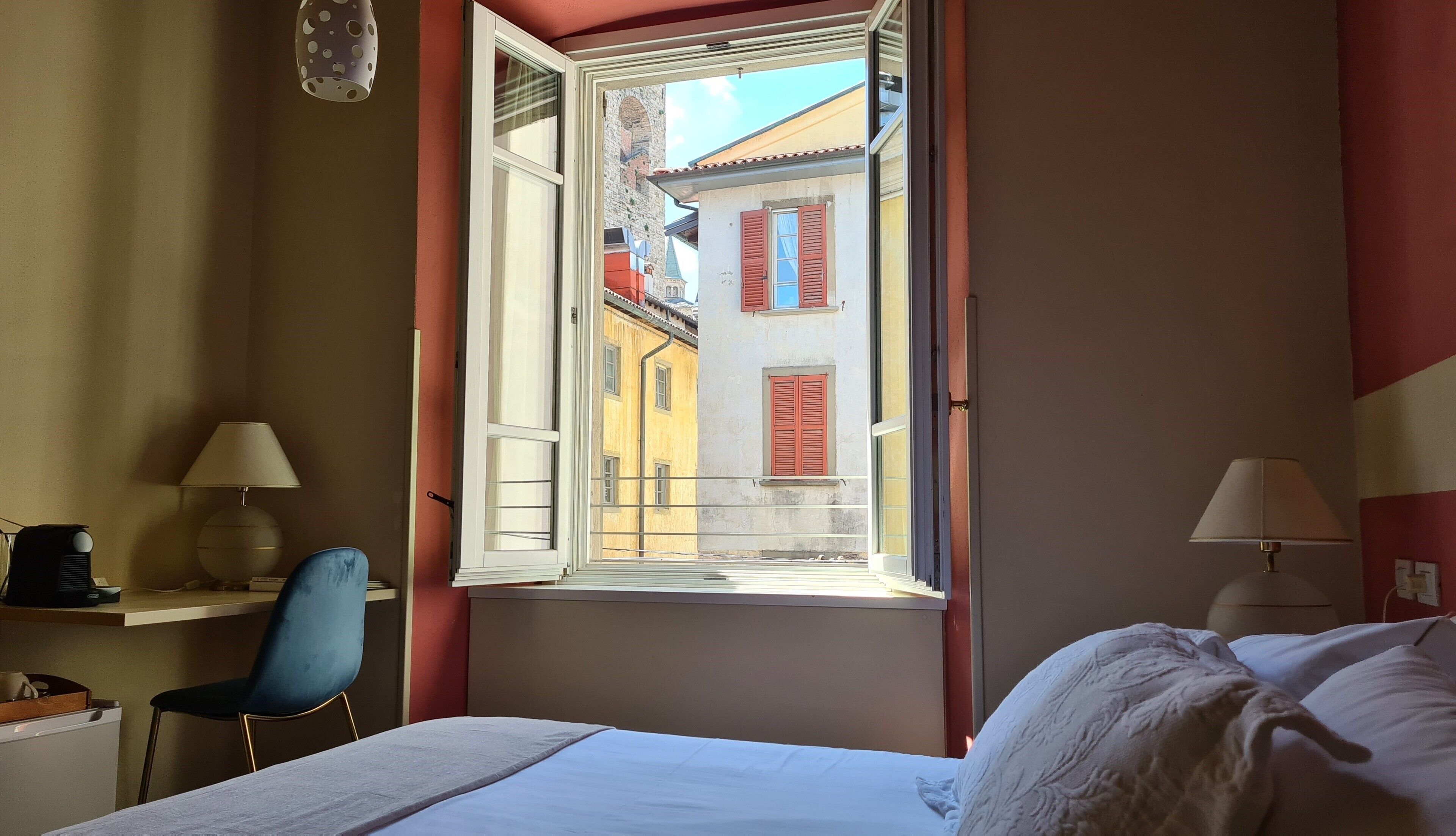 A hotel room with a bed, a desk with a chair, a window with red shutters, and a view of a building with a tower.