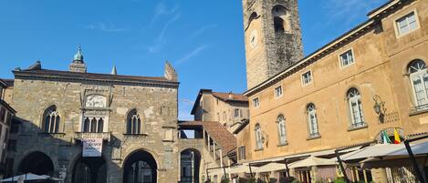 A historic square with a prominent tower, stone buildings, and outdoor seating areas.