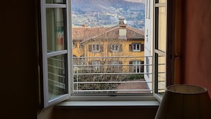 A room with a window overlooking a mountainous landscape, a lamp on a bedside table, and a view of a building with a tiled roof. - Hotel Piazza Vecchia (Bergamo)