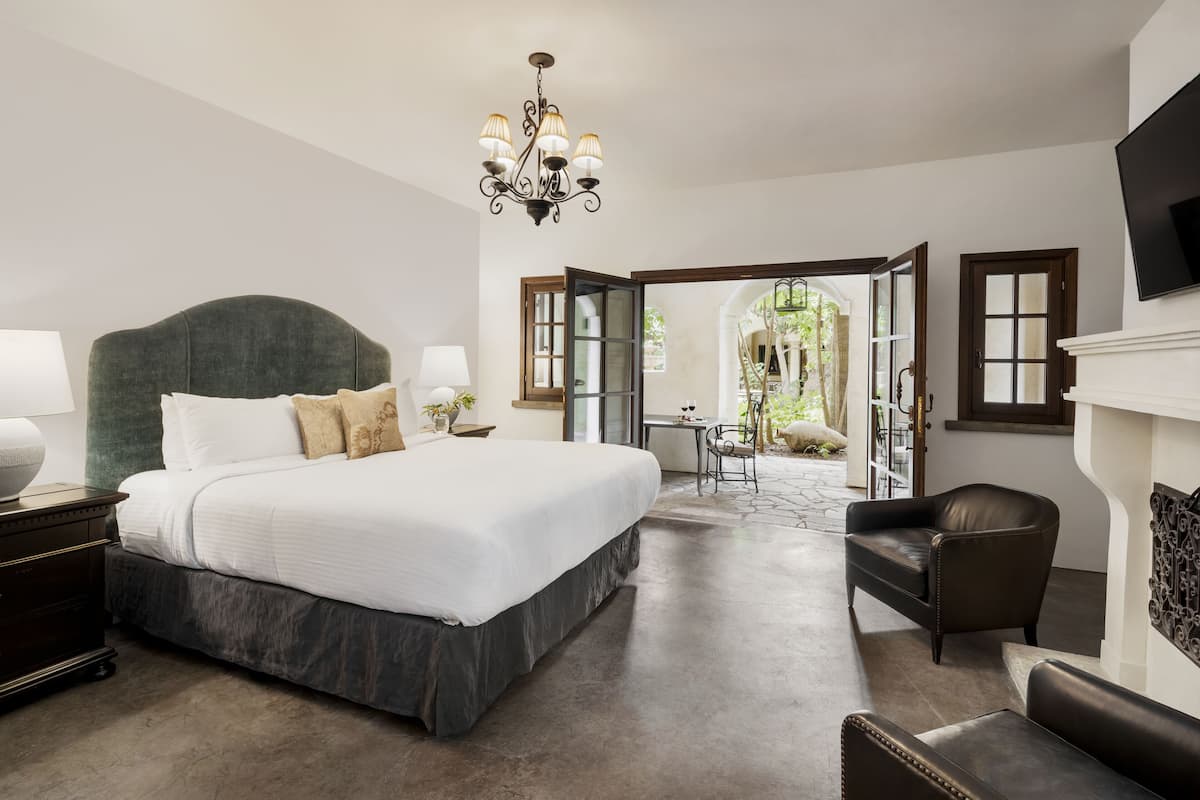 A spacious bedroom with a large bed, a chandelier, a fireplace, and an open door leading to a balcony.