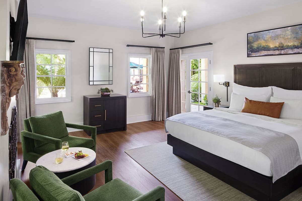 A modern bedroom with a large bed, green armchairs, a round table with a plate of fruit and a glass, and a view of the outdoors through a window.