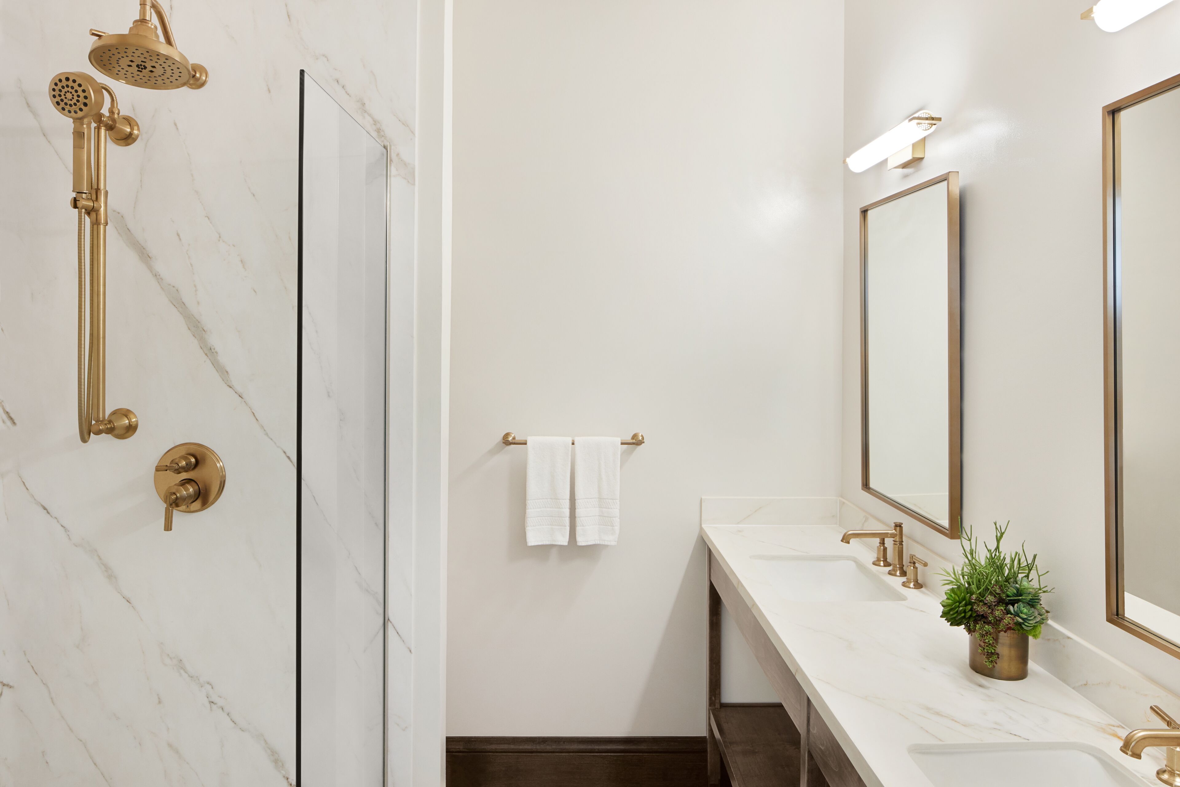 A modern bathroom with a marble wall, a gold showerhead, a sink with a potted plant, and a towel rack.