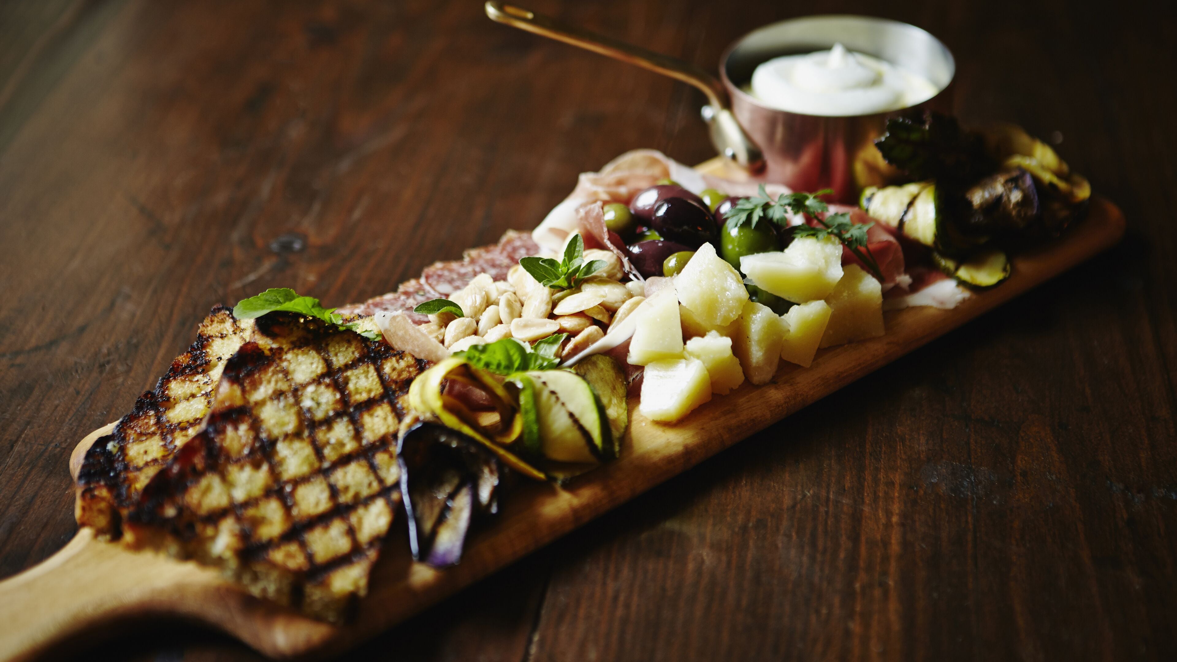 A wooden serving board with grilled cheese, olives, and a dipping sauce.