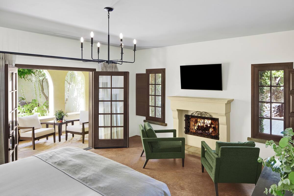A hotel room with a bed, two green armchairs, a fireplace, a TV mounted on the wall, and a view of the outdoors through large windows.