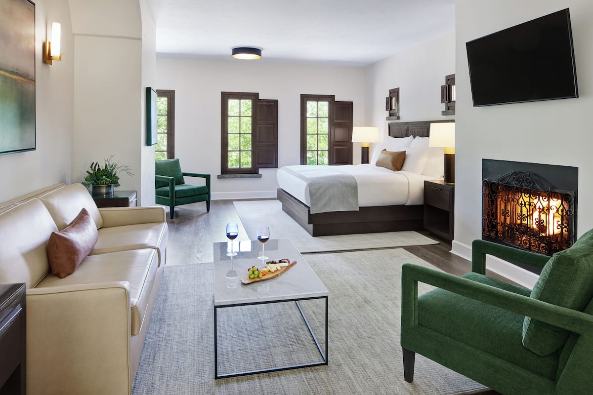 A modern hotel room with a bed, green armchairs, a coffee table with a fruit platter, and a fireplace.