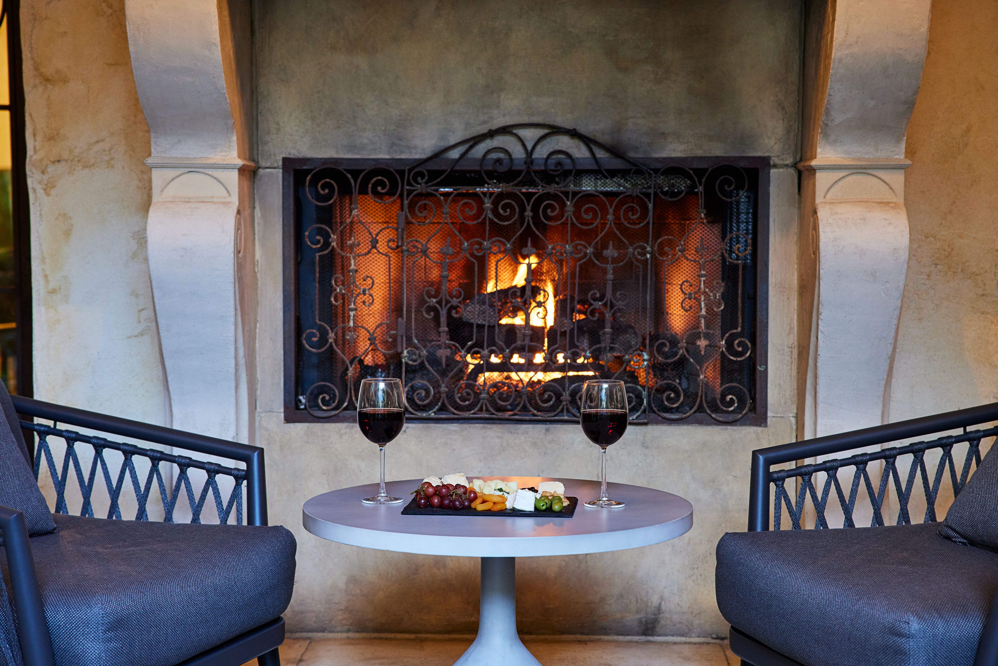 A cozy outdoor seating area with a fireplace, two chairs, a table with a cheese board and grapes, and two glasses of red wine.