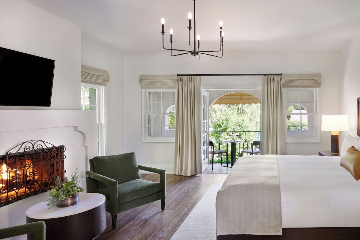A hotel room with a large bed, a green armchair, a small table with a plant, a fireplace, and a balcony with a view of greenery.
