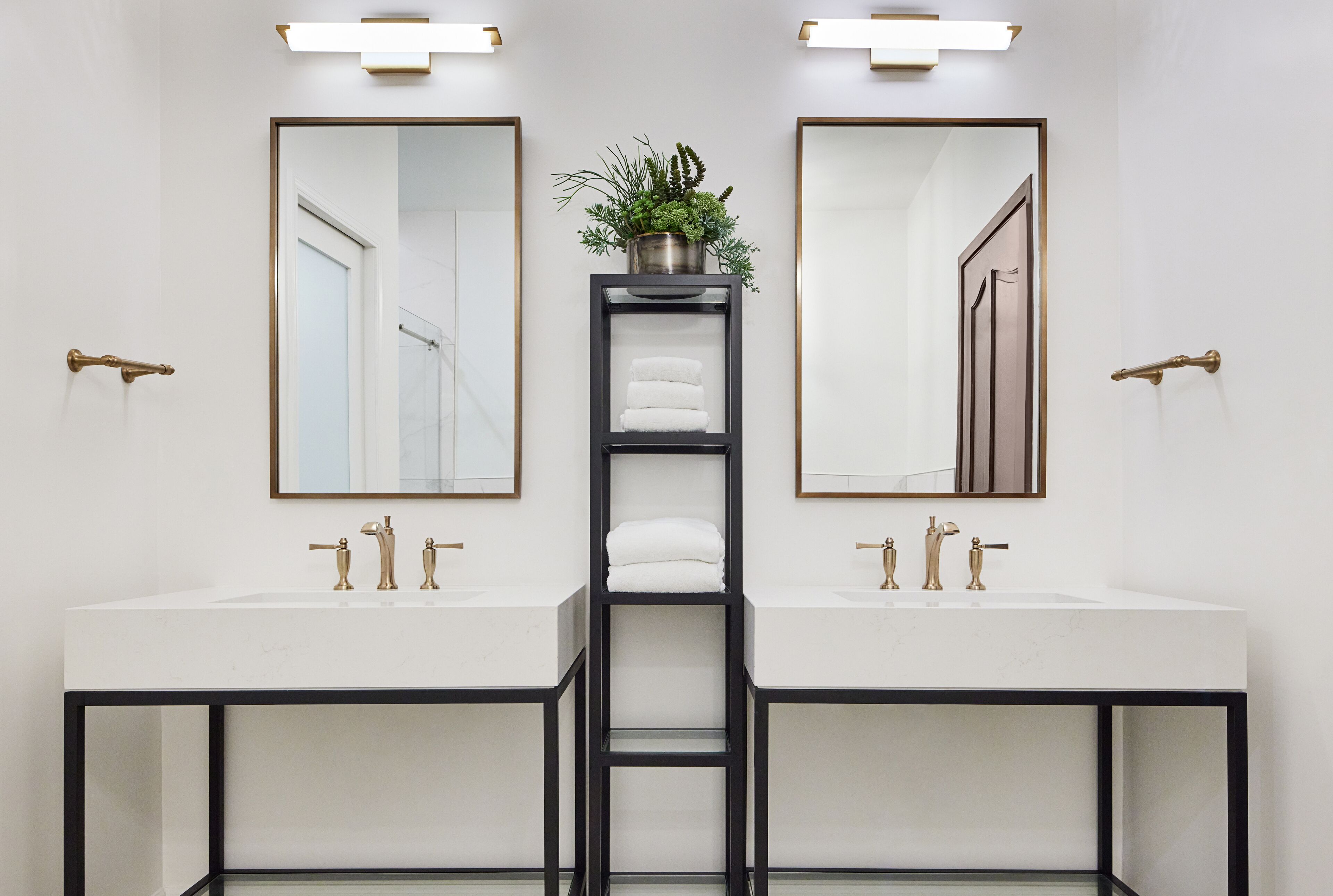 A modern bathroom with two sinks, a central shelf with towels, and a potted plant.