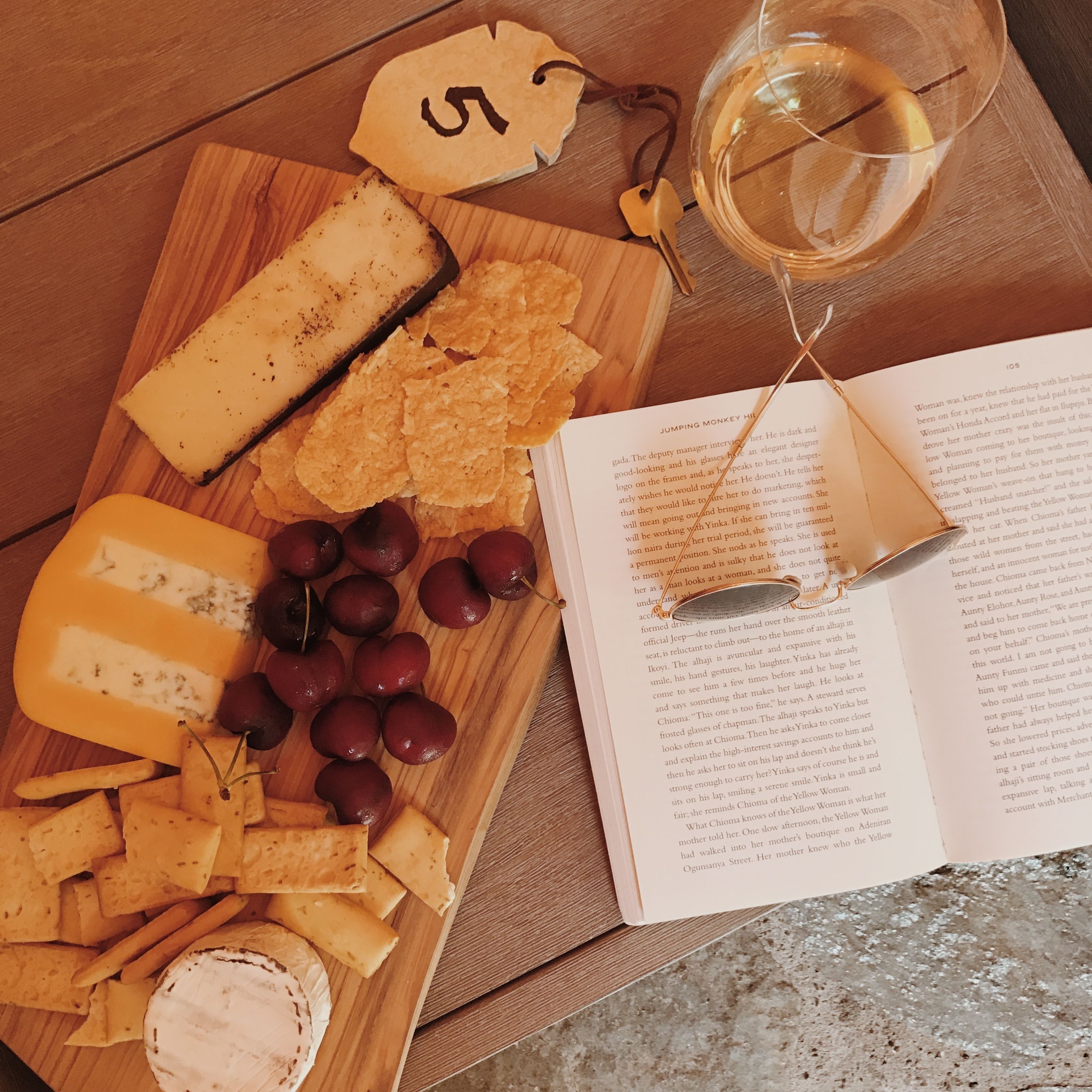 Una tabla de madera con diversos quesos, uvas y galletas, acompañada de un libro y una copa de vino.