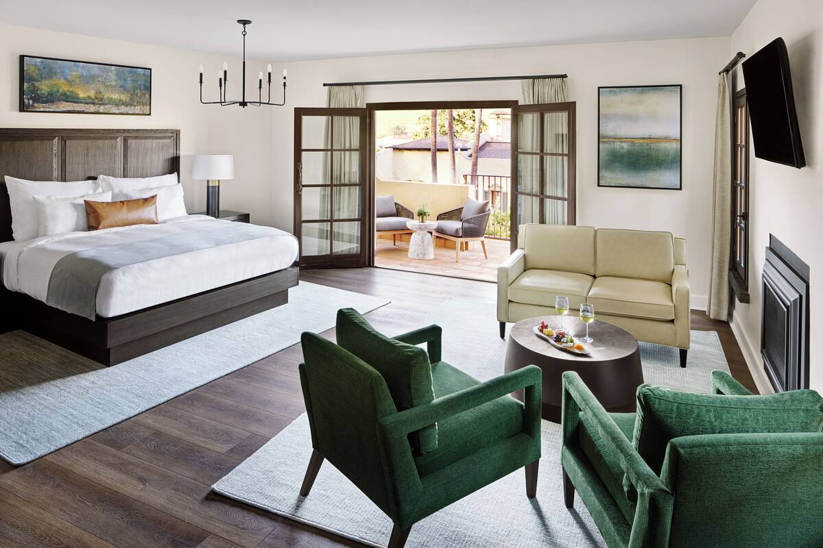 A hotel bedroom with a large bed, green armchairs, a sofa, a small table with drinks, and a balcony with outdoor seating.
