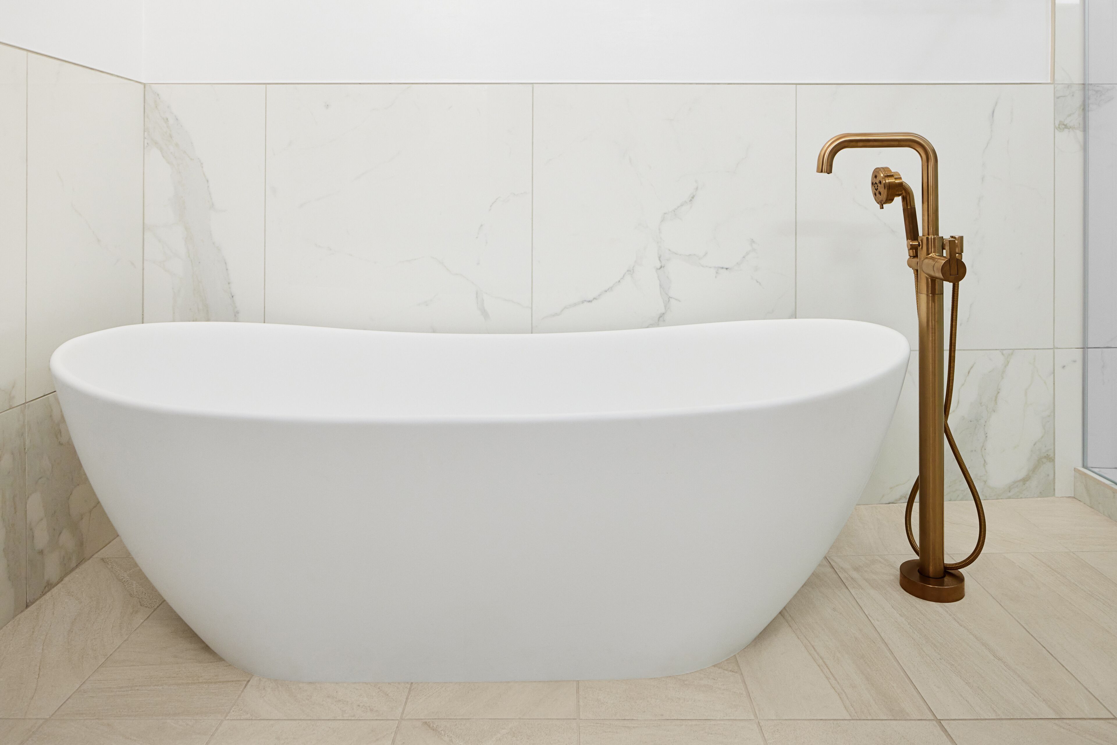 A freestanding bathtub with a gold faucet in a modern bathroom with marble walls and floor tiles.
