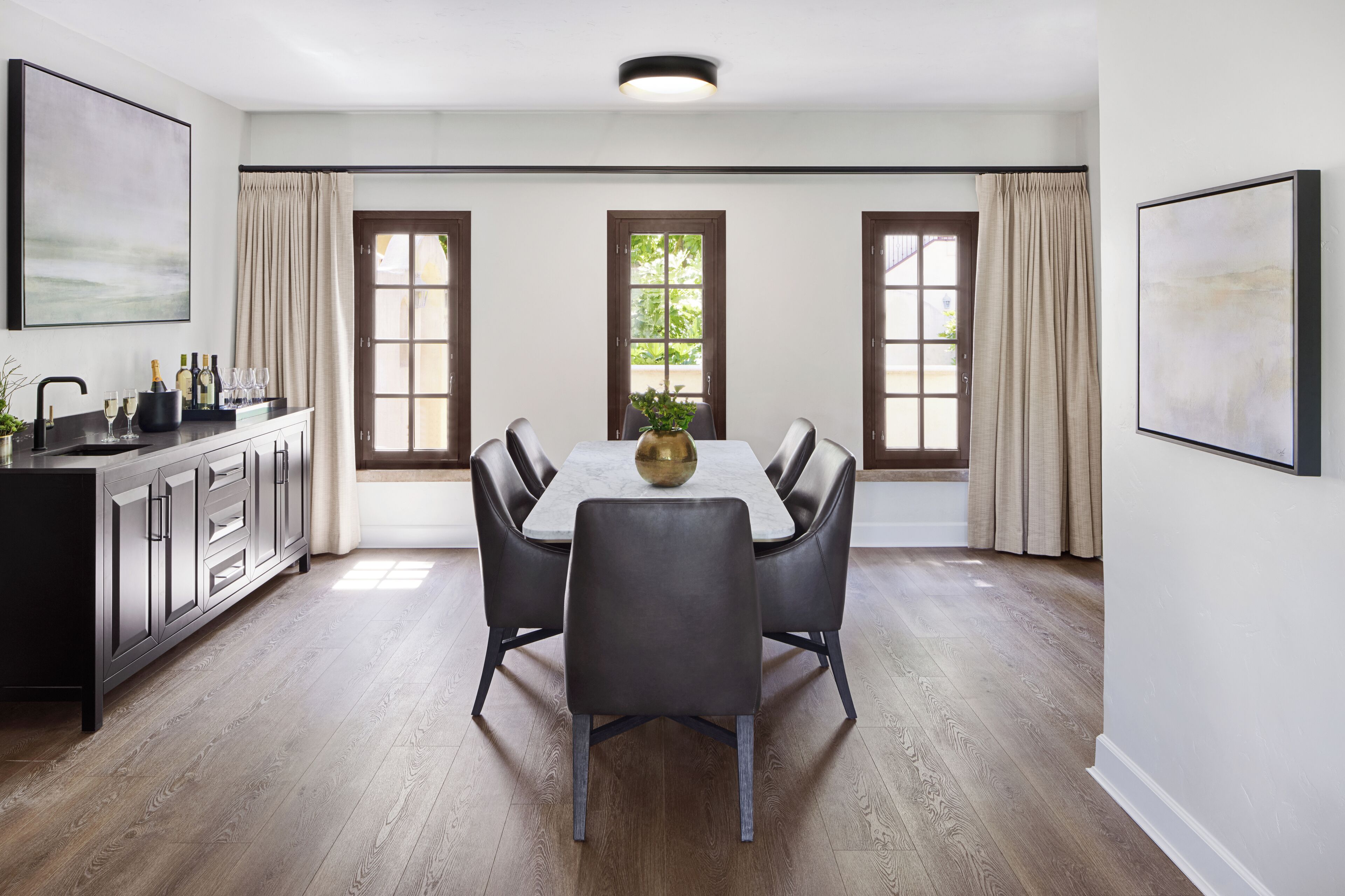 A dining area with a rectangular table, six chairs, a central vase, and a kitchen counter with cabinets and a sink.