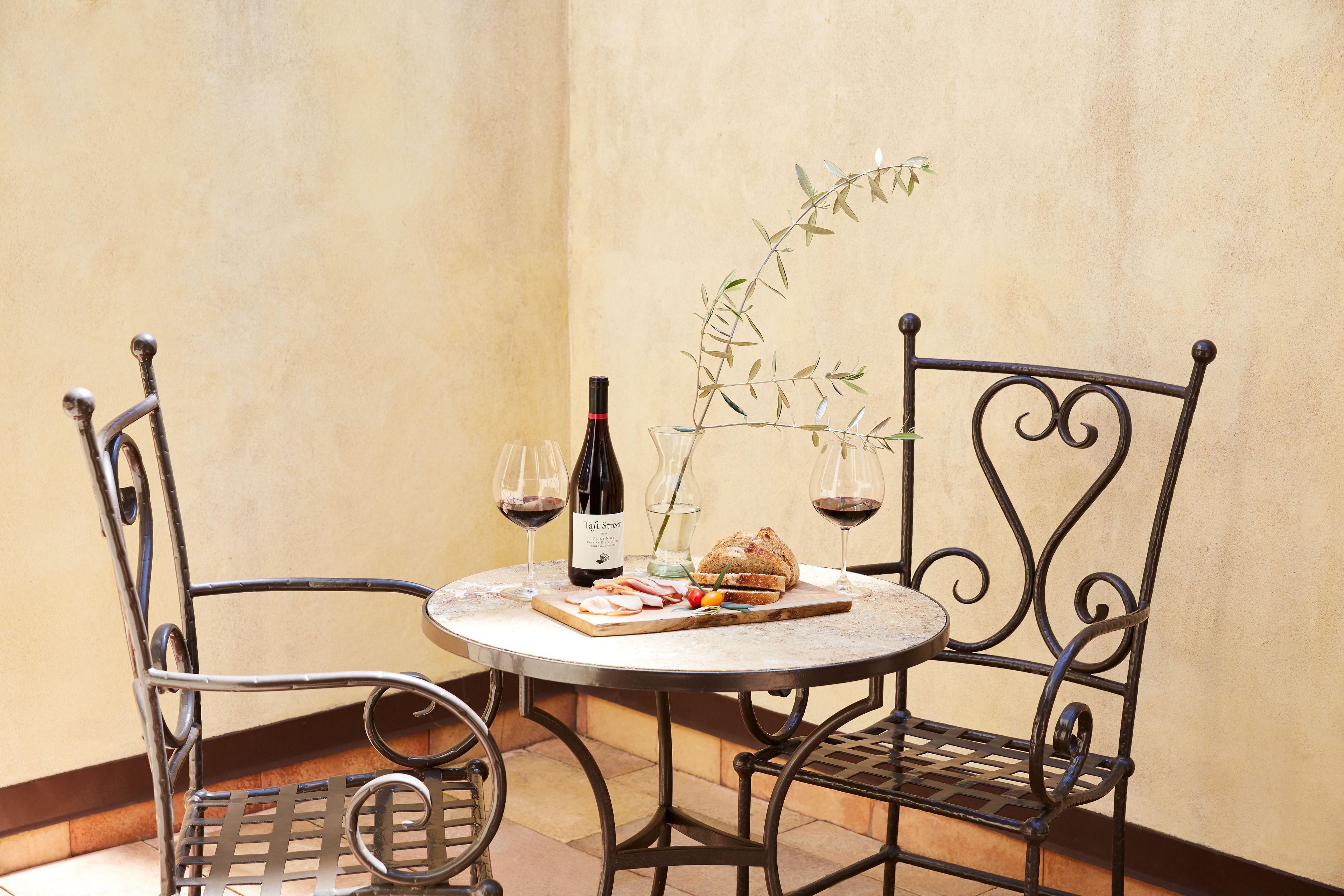 A round metal table with two chairs, a bottle of wine, a glass, a loaf of bread, and a small plant.