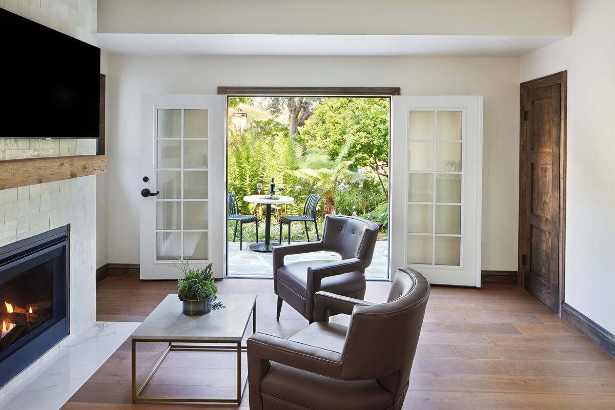 A living room with a fireplace, a coffee table, two leather chairs, and a view through glass doors to an outdoor patio with a table and chairs.