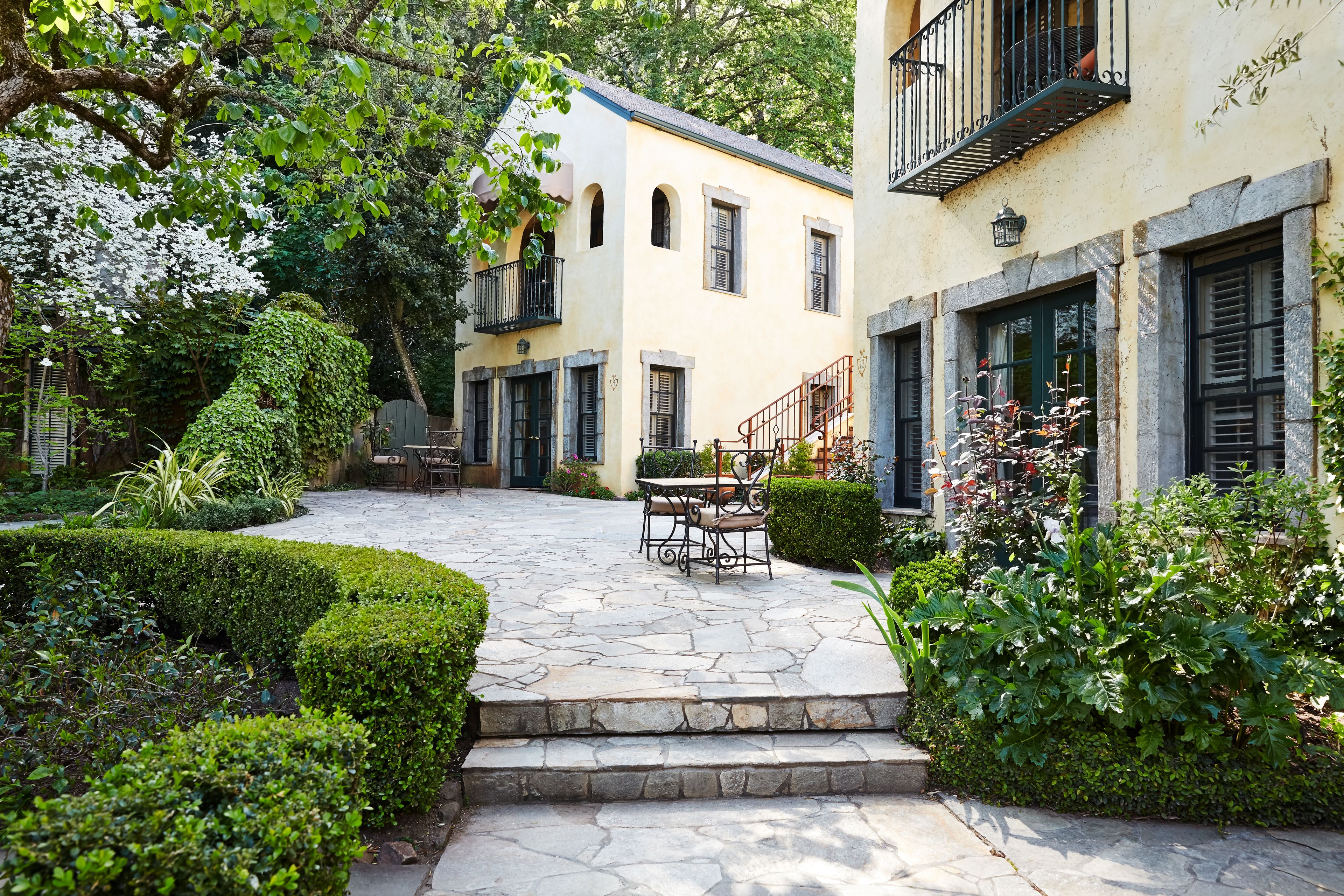 A courtyard with stone steps leading to a table and chairs, surrounded by well-maintained hedges and plants.