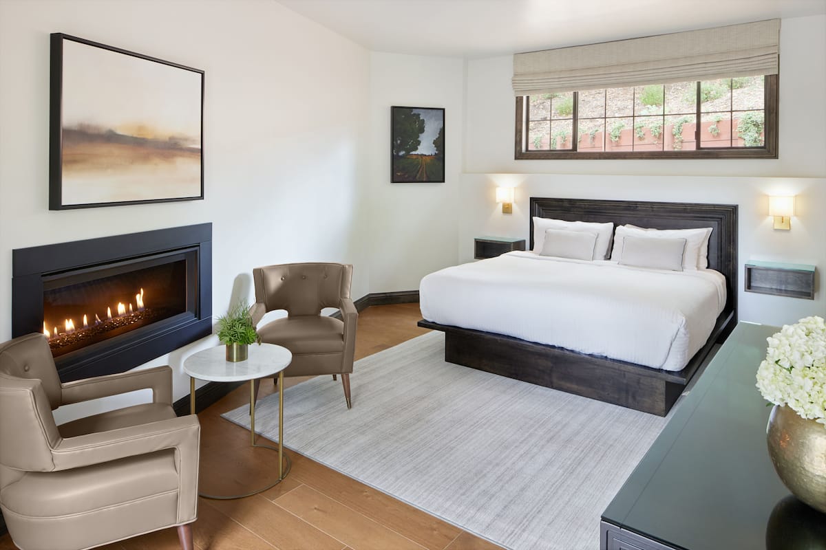 A bedroom with a fireplace, a bed, a chair, a small table, and a window with a view of greenery.