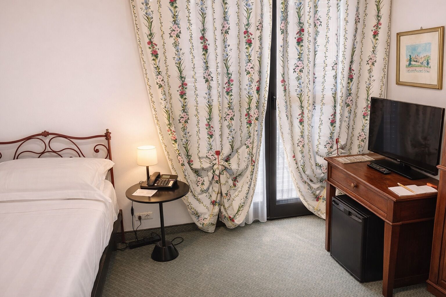 Camera d'albergo con un letto, una scrivania con televisore, una lampada e un telefono.