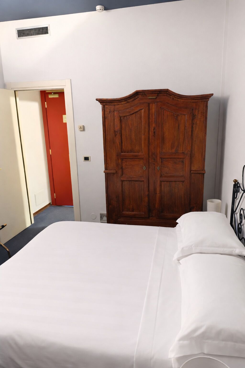 A bedroom with a large wooden wardrobe, a bed with white linens, and a red door in the background.