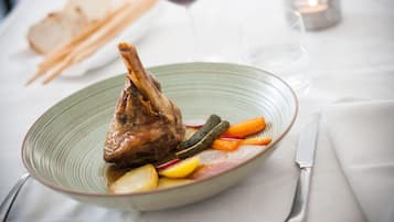 A plate with a lamb shank, vegetables, and a glass of red wine on a table.