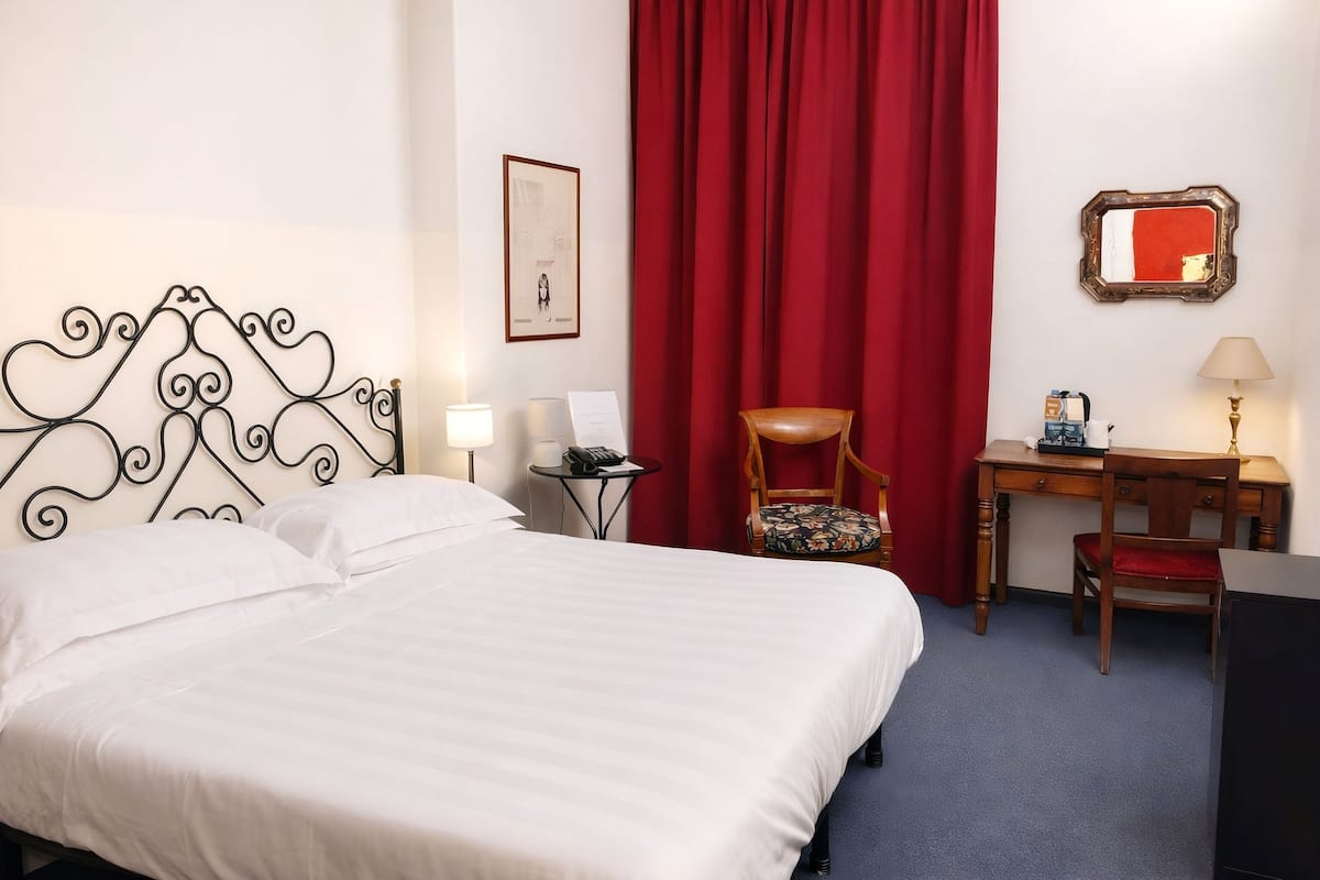 A hotel room with a bed, a desk, a chair, a lamp, and a red curtain.