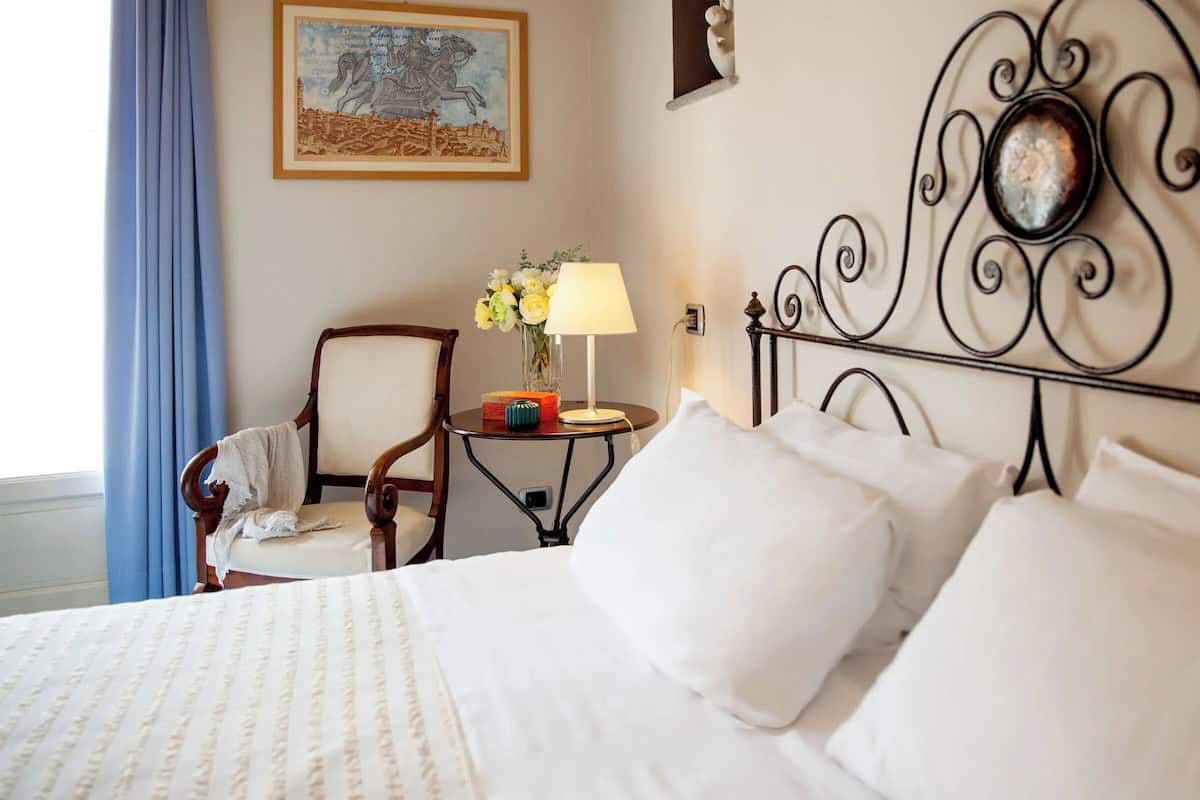 A neatly made bed with white linens, a wooden chair, a small table with a lamp and flowers, and a framed artwork on the wall.