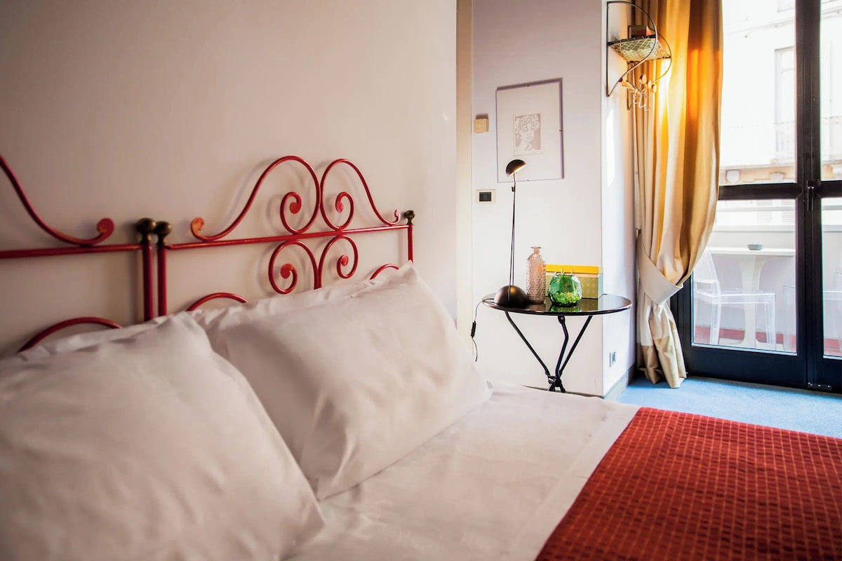 A hotel room with a red metal bed frame, white bedding, a small side table with a lamp, and a view of the outside through a window with curtains.