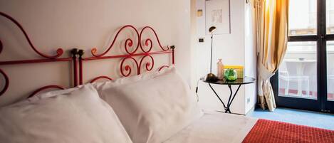 A hotel room with a red metal bed frame, white bedding, a small side table with a lamp, and a view of the outside through a window with curtains.