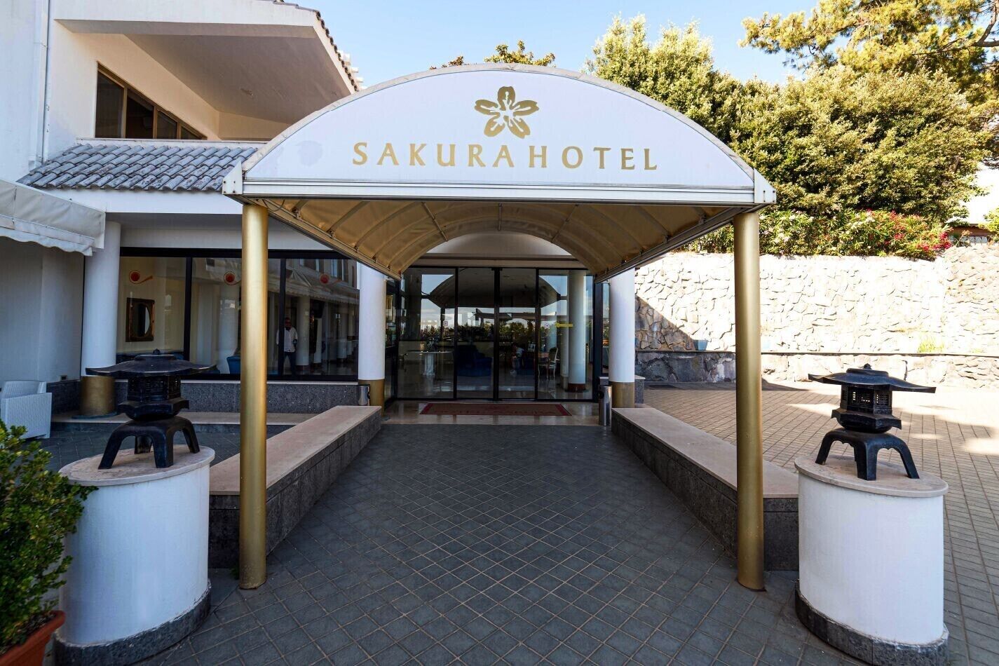 The entrance to the Sakura Hotel with a covered walkway, two lanterns, and a stone wall in the background.