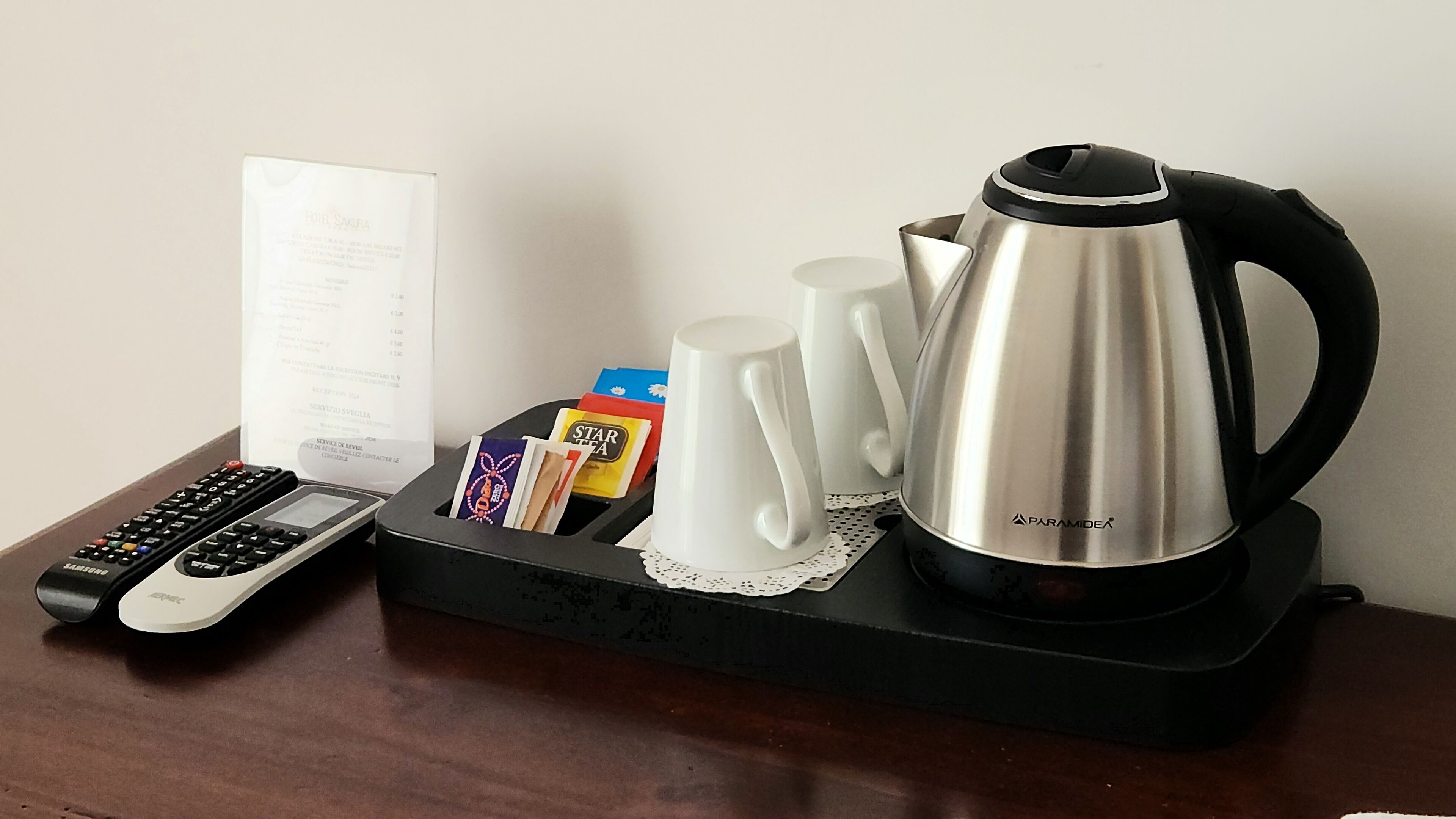 A tray with a remote control, a menu, a coffee pot, and some cups and a box of coffee on a wooden table.