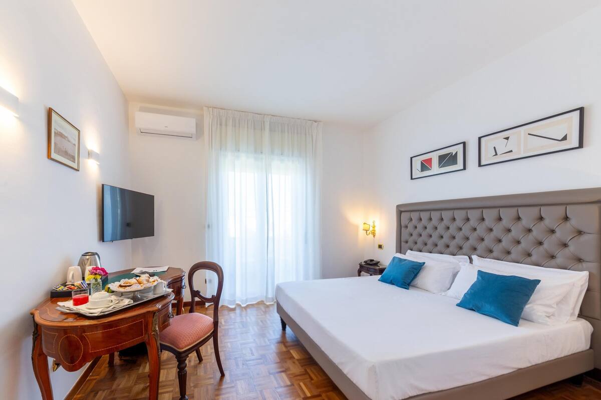 A hotel room with a bed, a desk with breakfast, a TV, and artwork on the walls.