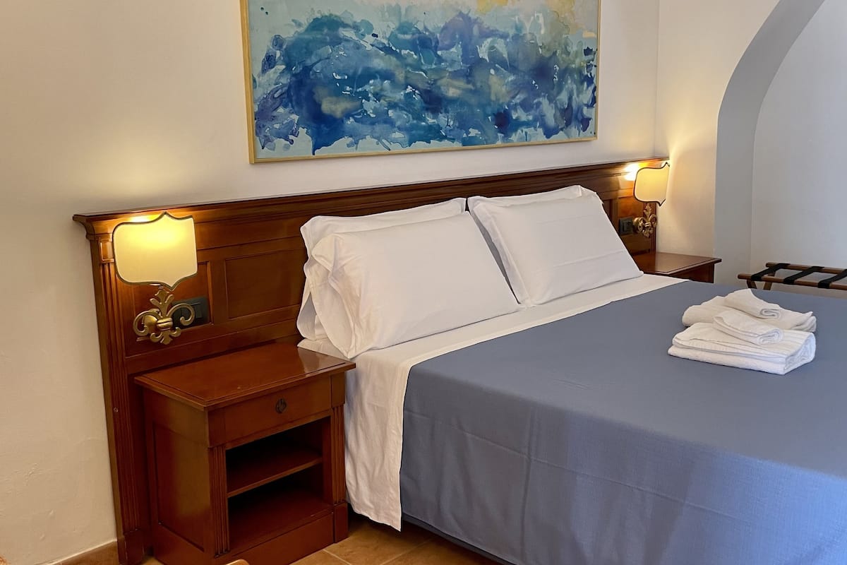 Een net opgemaakt bed met witte lakens, een blauwe sprei en een opgevouwen handdoek aan de zijkant. Er staat een houten nachtkastje met een lamp, een schilderij aan de muur en een houten stoel.
