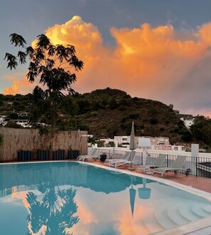 Una piscina con sillones de descanso, una cerca de madera y vistas a una colina con edificios al fondo.
