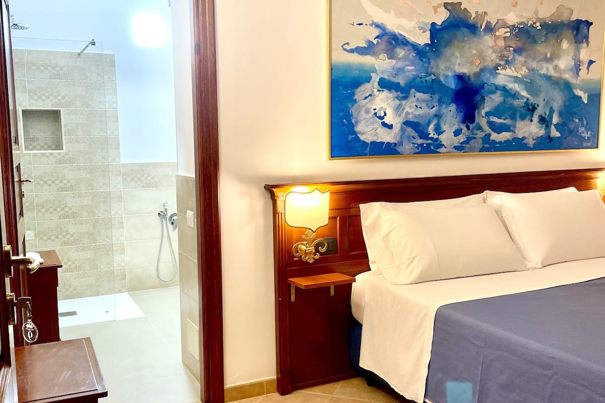 Een hotelkamer met een bed, nachtkastje, een wandlamp, een schilderij en uitzicht buiten via een glazen deur.