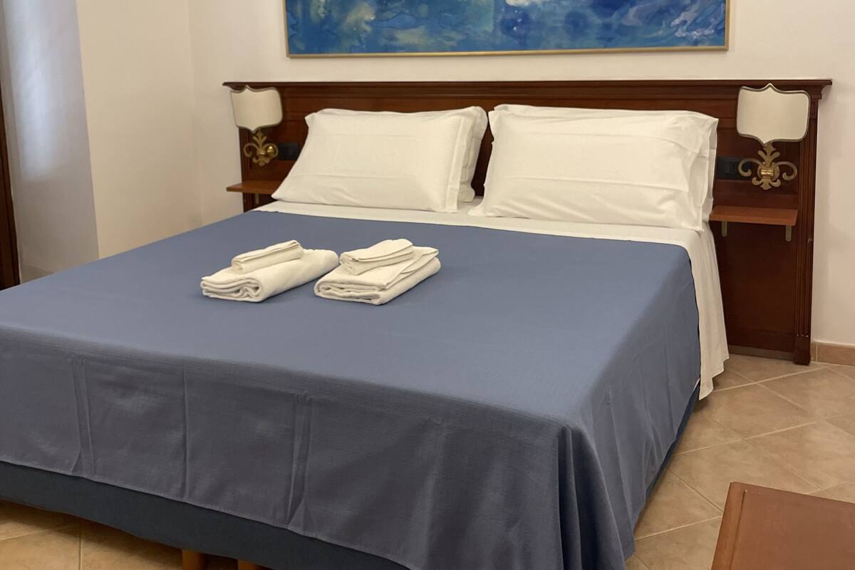 Een keurig opgemaakt bed met een blauwe dekbedovertrek en witte kussens, een schilderij boven het bed en een klein tafeltje ernaast.