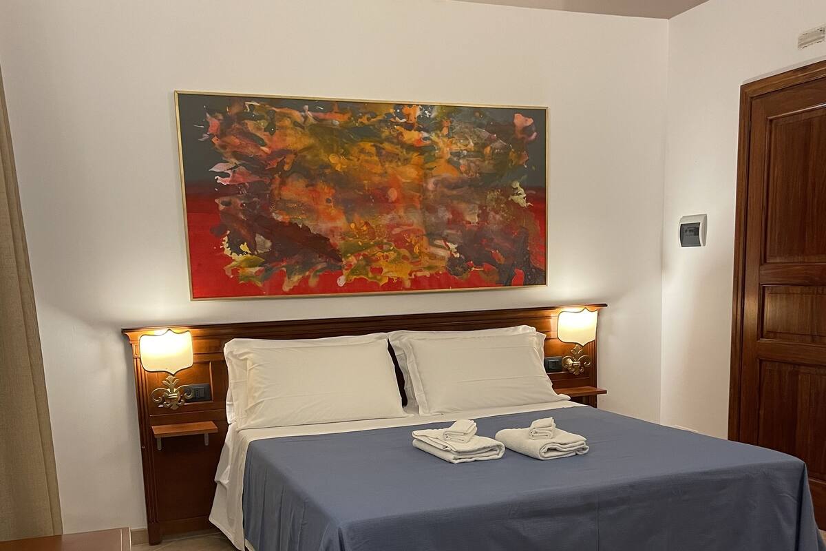 Een hotelkamer met een bed, twee nachtkastjes, een houten deur en een groot abstract schilderij aan de muur.