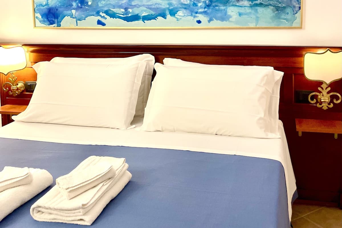 Een hotelkamer met een groot abstract schilderij boven het bed, een opgemaakt bed met witte lakens en een blauwe sprei.