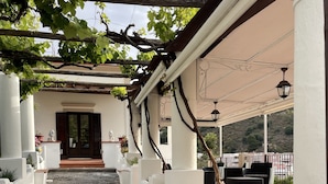 Un paseo techado al aire libre que conduce a la entrada de un edificio, con una pérgola, plantas en macetas y mobiliario exterior.