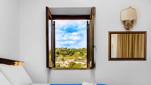 Una habitación de hotel con una cama, un ventanal que ofrece vistas a un pueblo, una lámpara fijada a la pared y un espejo enmarco.