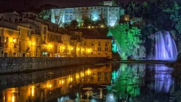 A nighttime view of a historic town with illuminated buildings, a calm river reflecting the lights, and a waterfall in the background.