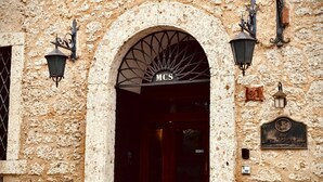 An arched entrance to a building with a sign reading "MCS" above the door. There are two potted plants on either side of the entrance, and a plaque on the wall to the right.