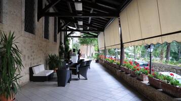 A covered outdoor seating area with a sofa and chairs, potted plants, and a view of the street.