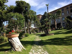 A courtyard with a stone pathway, lush greenery, palm trees, and a building with multiple windows. - Hotel Il Cavalier D'Arpino (Arpino)