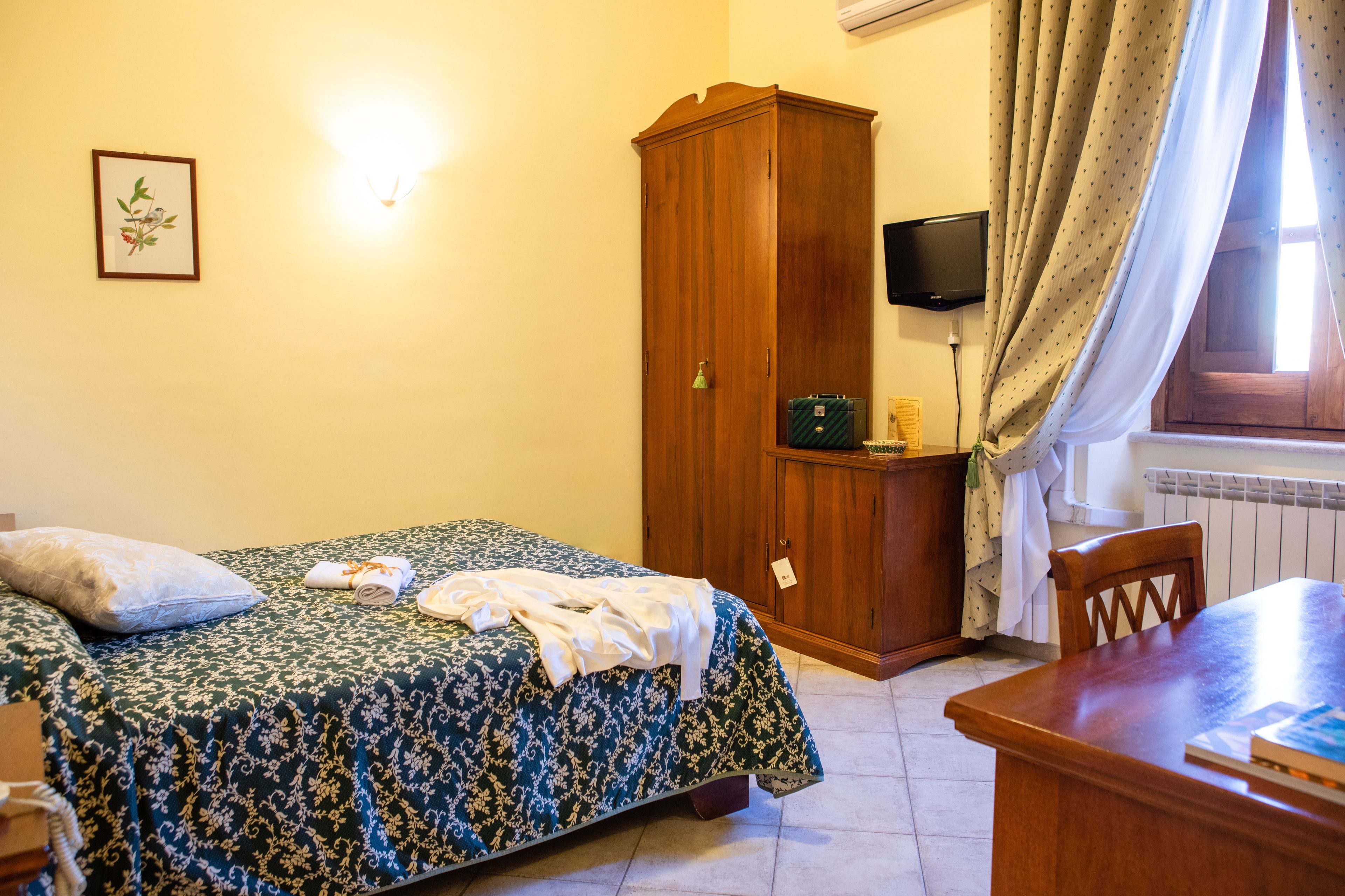 Une chambre à coucher comprenant un lit, une armoire en bois, un bureau, une chaise, un téléviseur et une fenêtre avec des rideaux.