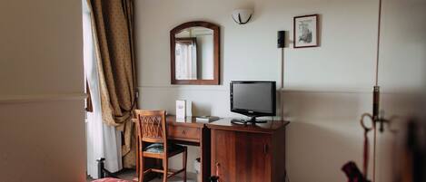 A hotel room with a bed, a desk with a chair, a television, a wardrobe, a mirror, and an air conditioning unit.