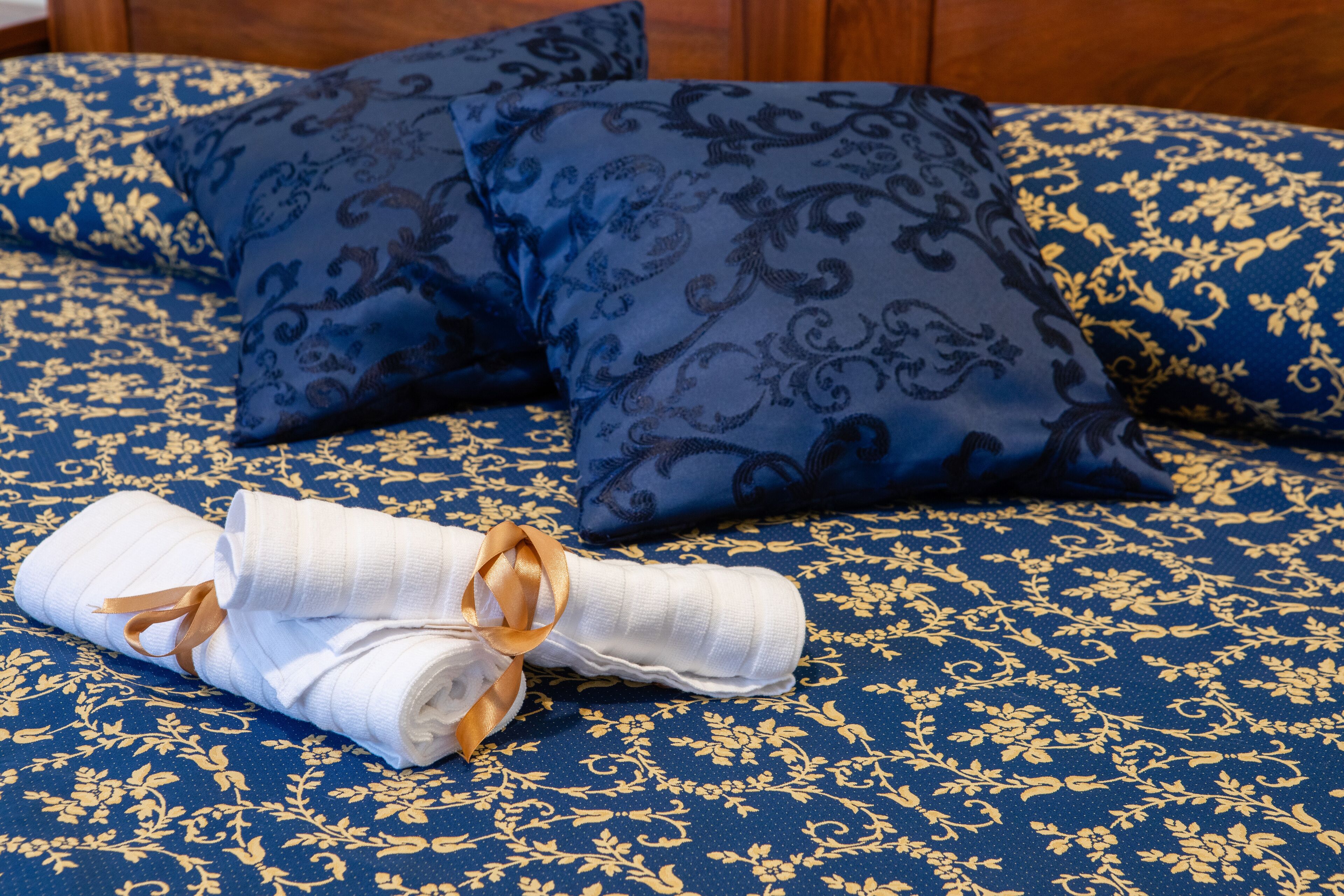 Una cama con ropa de cama de diseño azul y dorado, una toalla blanca doblada con un lazo dorado, y dos almohadas a juego.