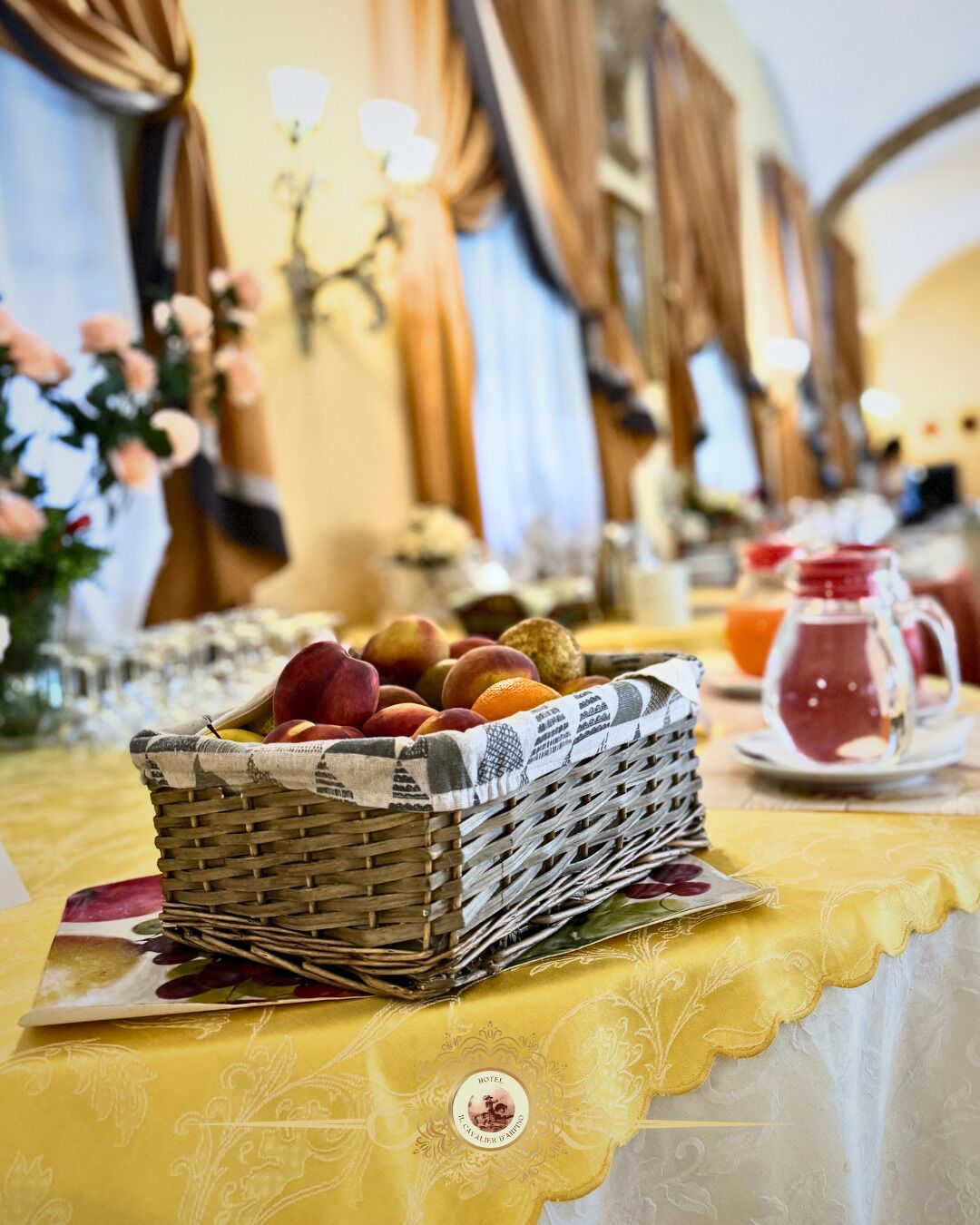 A dining table with a wicker basket of fruits, a yellow tablecloth, and a pitcher of red beverage.