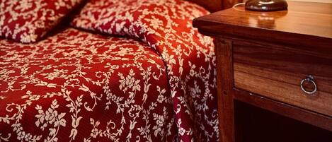 A hotel room with a wooden headboard, a red patterned bedspread, and a wooden nightstand with a lamp.