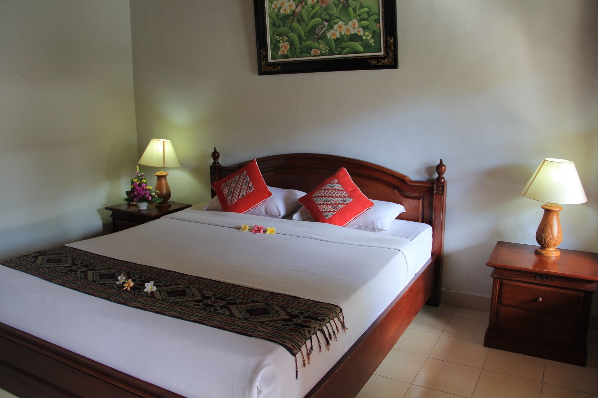 A neatly made bed with white linens, a patterned bedspread, and red decorative pillows. There are two wooden nightstands with lamps, a framed picture on the wall, and a small table with a vase of flowers.