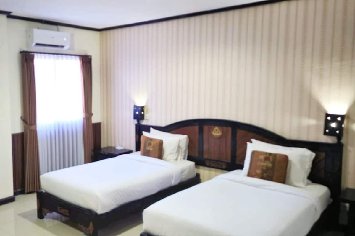 A hotel room with two single beds, a wardrobe, a window with curtains, an air conditioning unit, and a nightstand with a lamp.