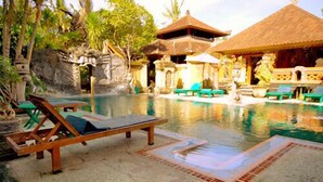 A pool area with lounge chairs, a stone structure, and a thatched-roof building. - Stana Puri Gopa Hotel (Denpasar)